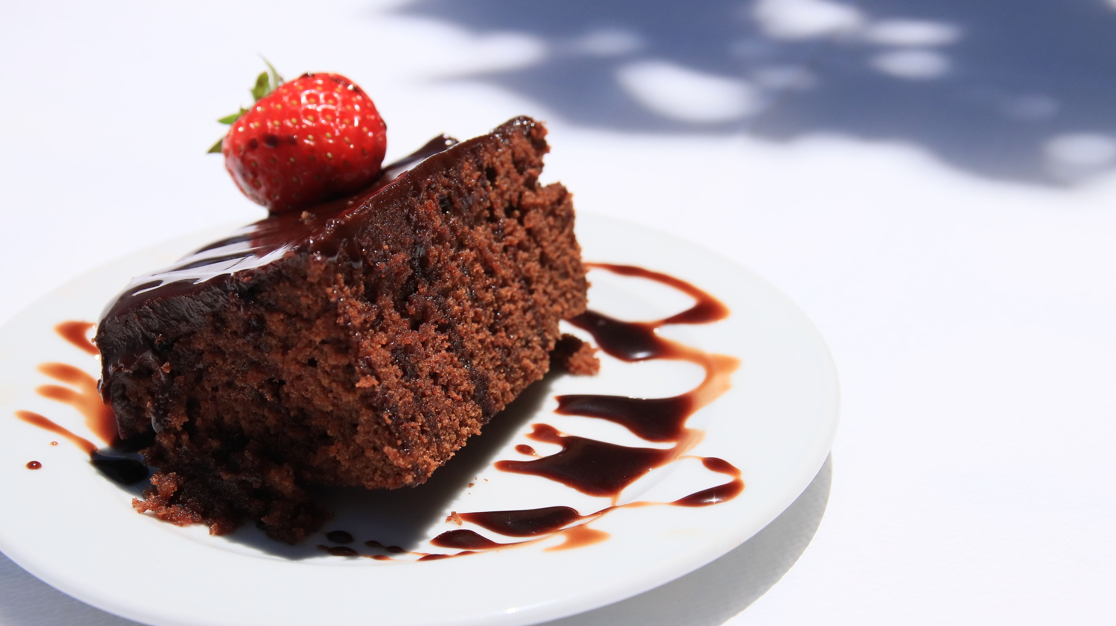Uma fatia de bolo de chocolate com uma morango por cima, servida em um prato branco com calda de chocolate.