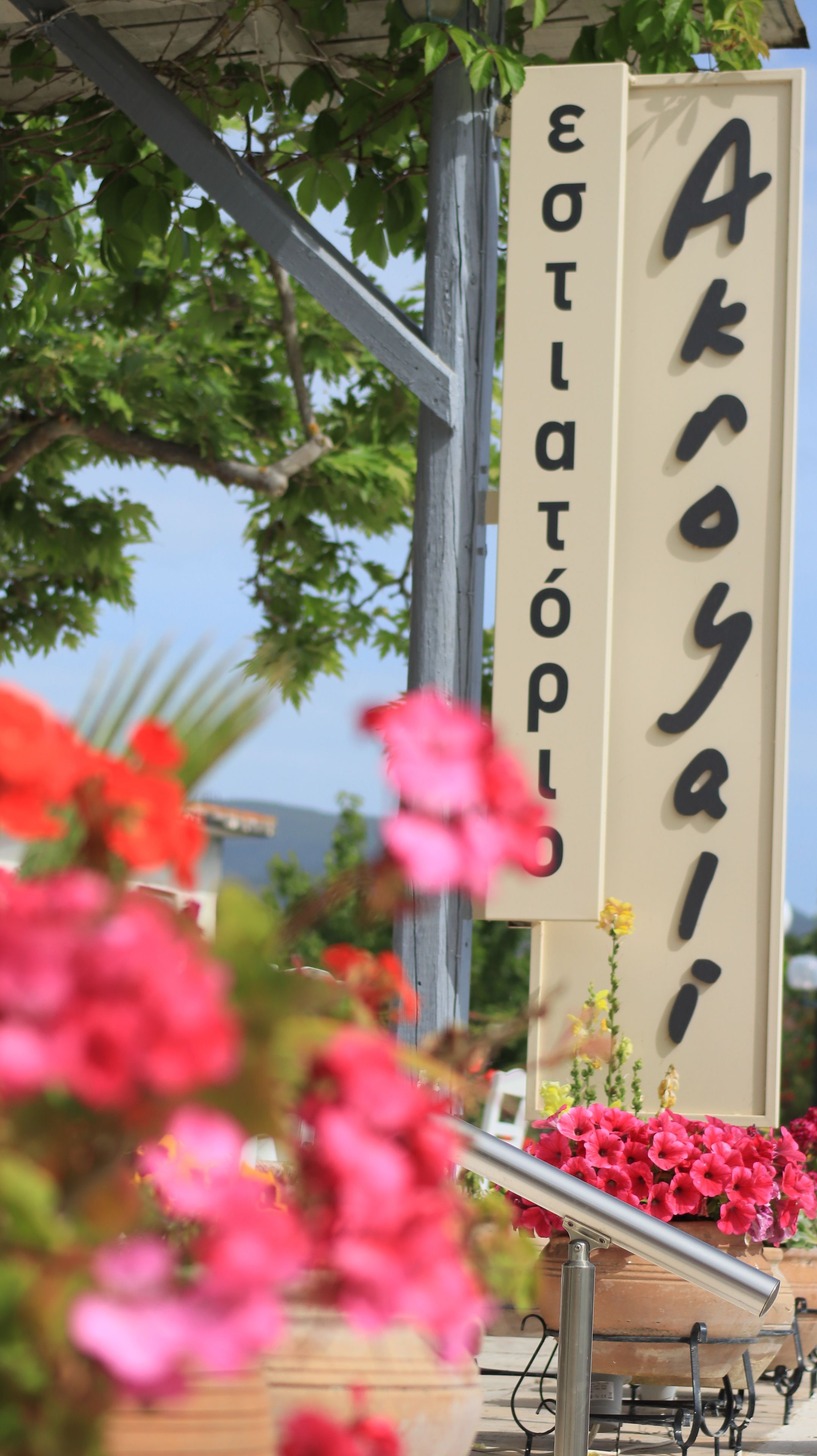 A sign with the text "Akroya" and "Eotatopoi" in a vertical layout, surrounded by greenery and flowers.