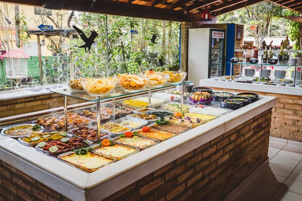 A buffet with various food items and a view of greenery outside. - Hotel Pousada Vovozinho (Guacui)