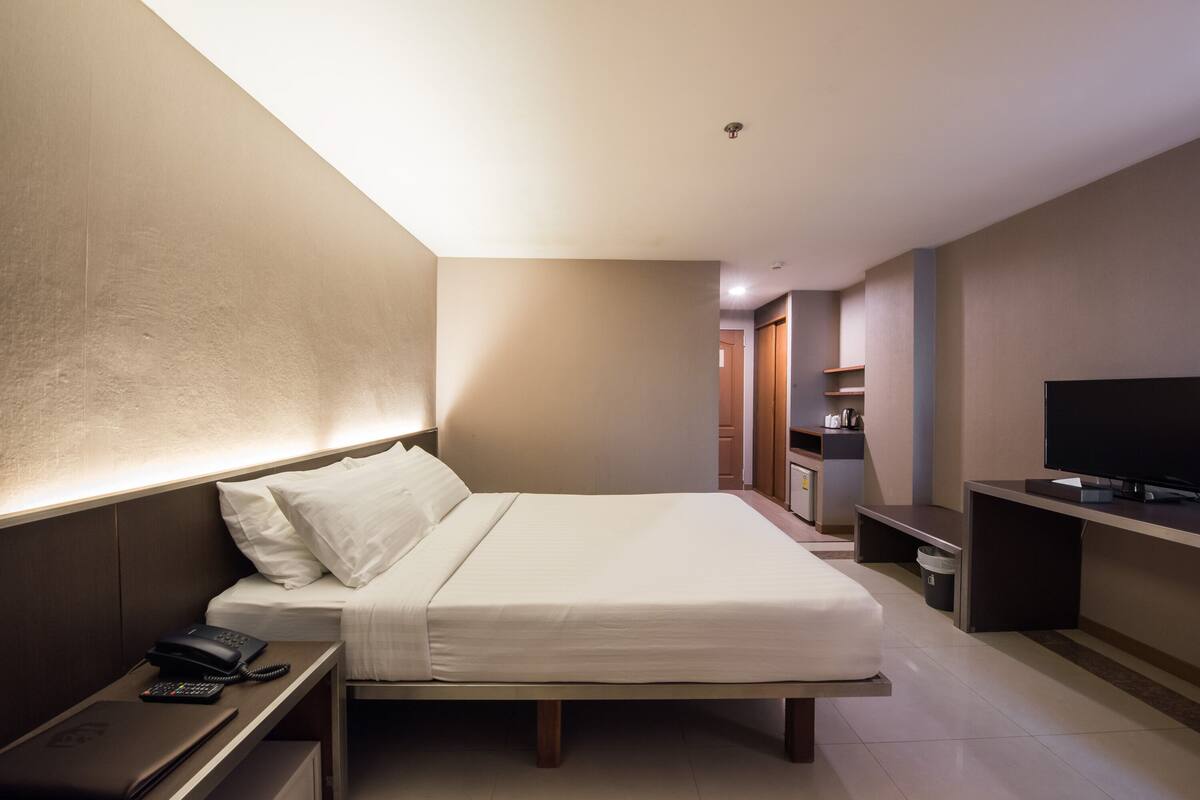 Quarto de hotel com cama, mesa de cabeceira, televisão e pequena escrivaninha com cadeira.