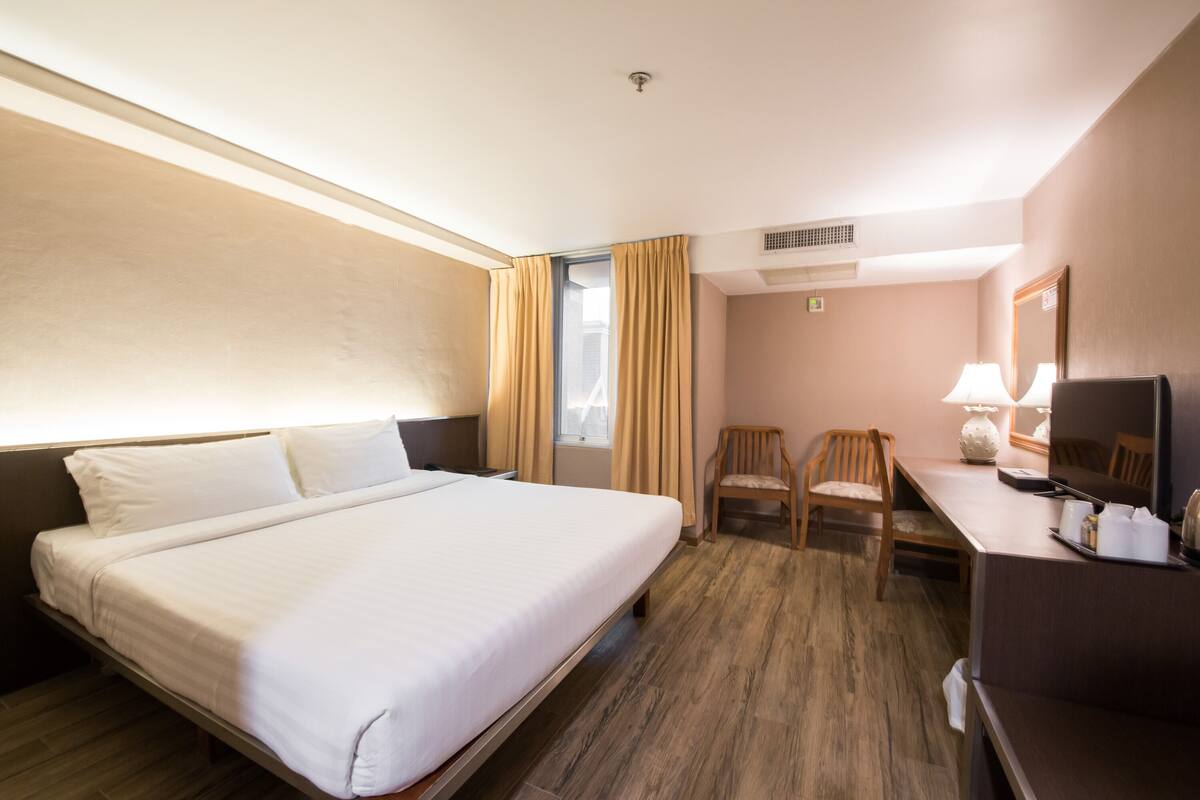 Quarto de hotel com uma cama grande, uma escrivaninha com cadeira, uma televisão e uma janela com cortinas.