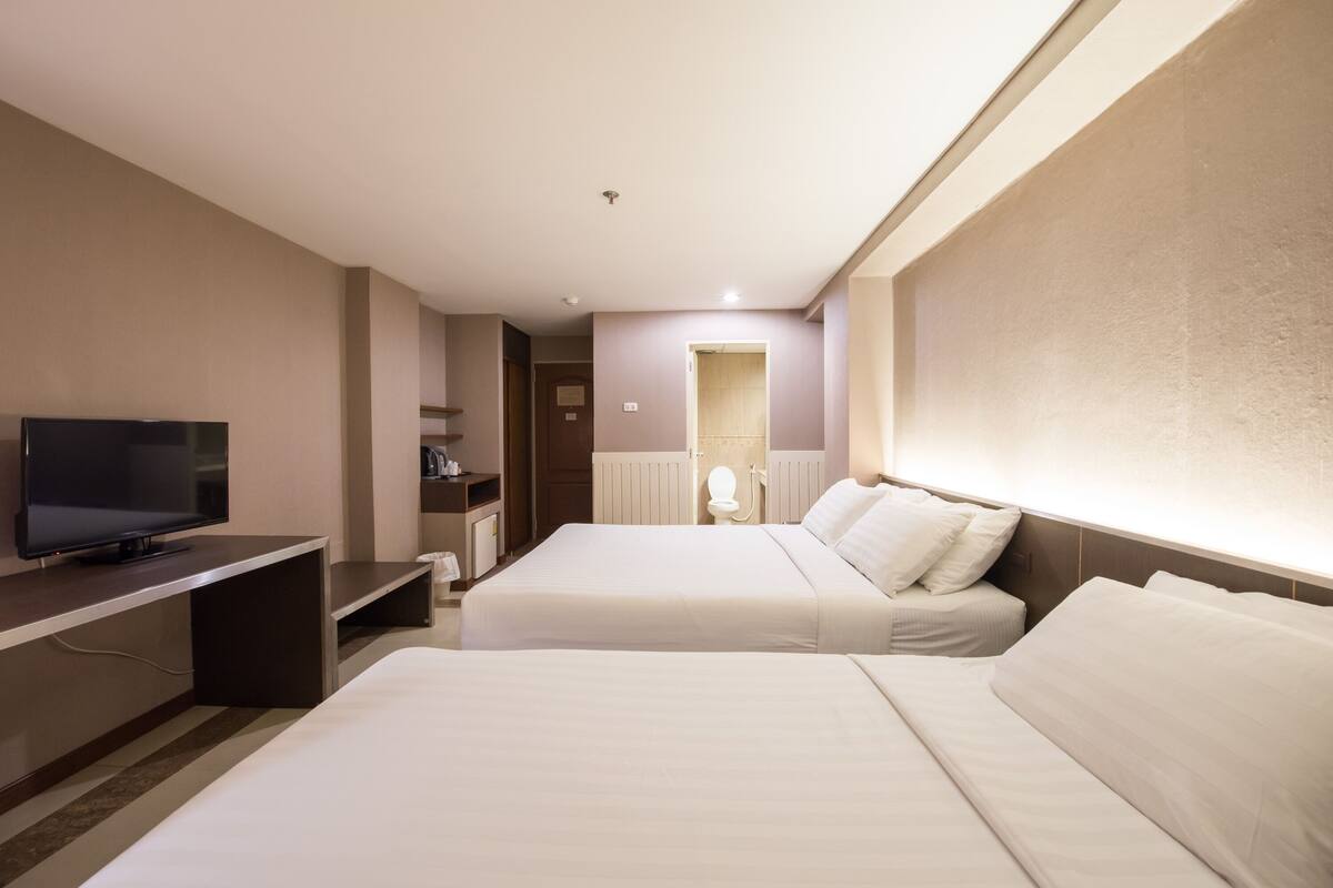 Quarto de hotel com duas camas, televisão, escrivaninha e banheiro ao fundo.