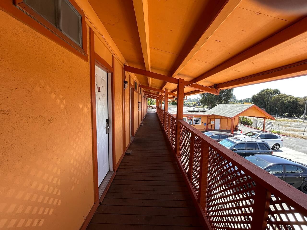 A covered corridor with wooden railings, a door on the left, and a view of parked cars and a building in the background.