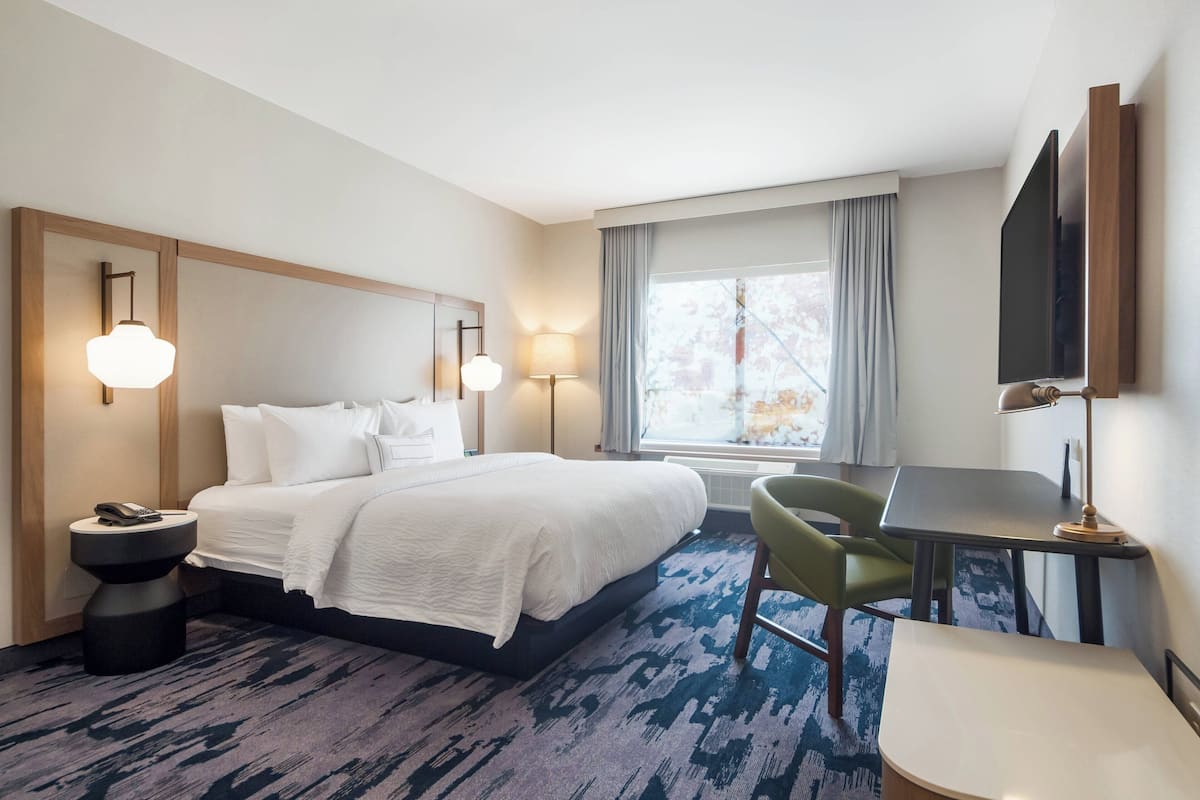 A hotel room with a large bed, two bedside lamps, a desk with a chair, a flat-screen TV, and a window with curtains.