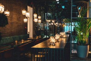 Un comedor interior con varias mesas y sillas, lámparas colgantes y una planta grande en maceta.