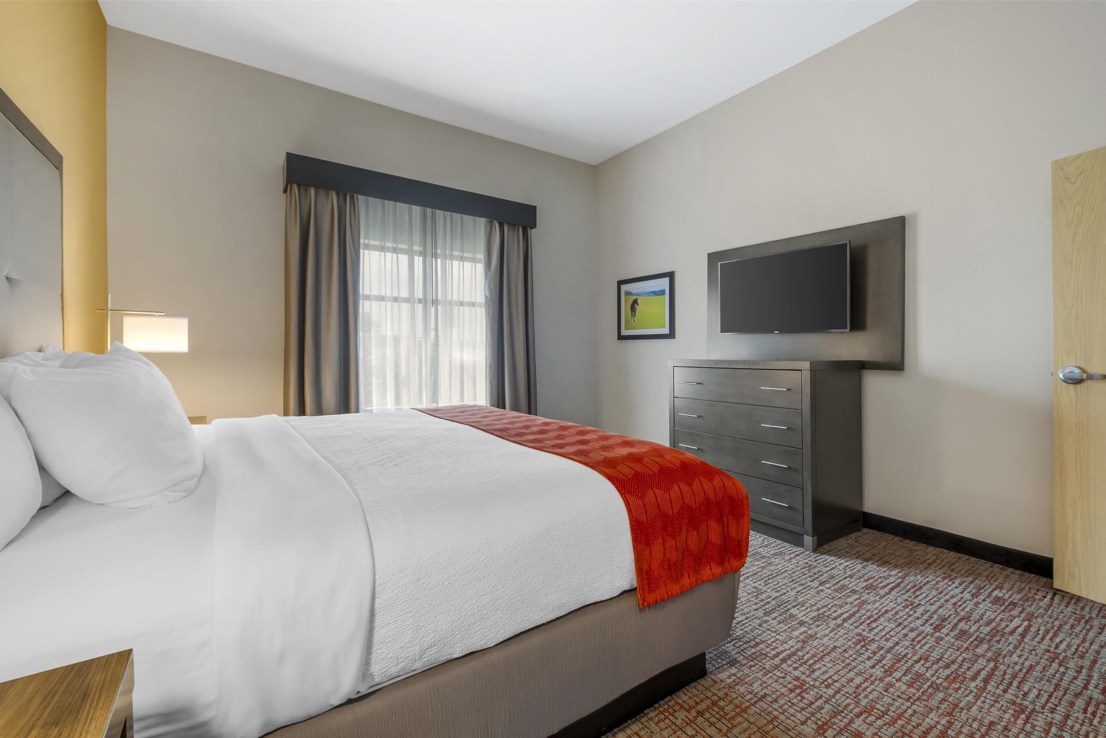 A hotel room with a bed, a television mounted on the wall, a dresser, and a window with curtains.