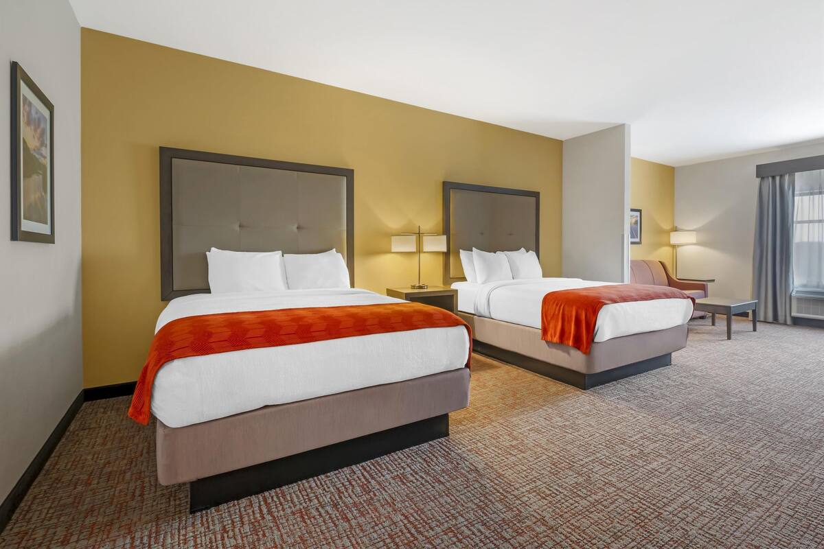 A hotel room with two beds, each with a headboard, white bedding, and an orange throw blanket. There are two bedside lamps and a small table with a chair.
