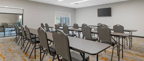 A conference room with multiple tables and chairs, a flat-screen TV on the wall, and a large window with a view of the outdoors.