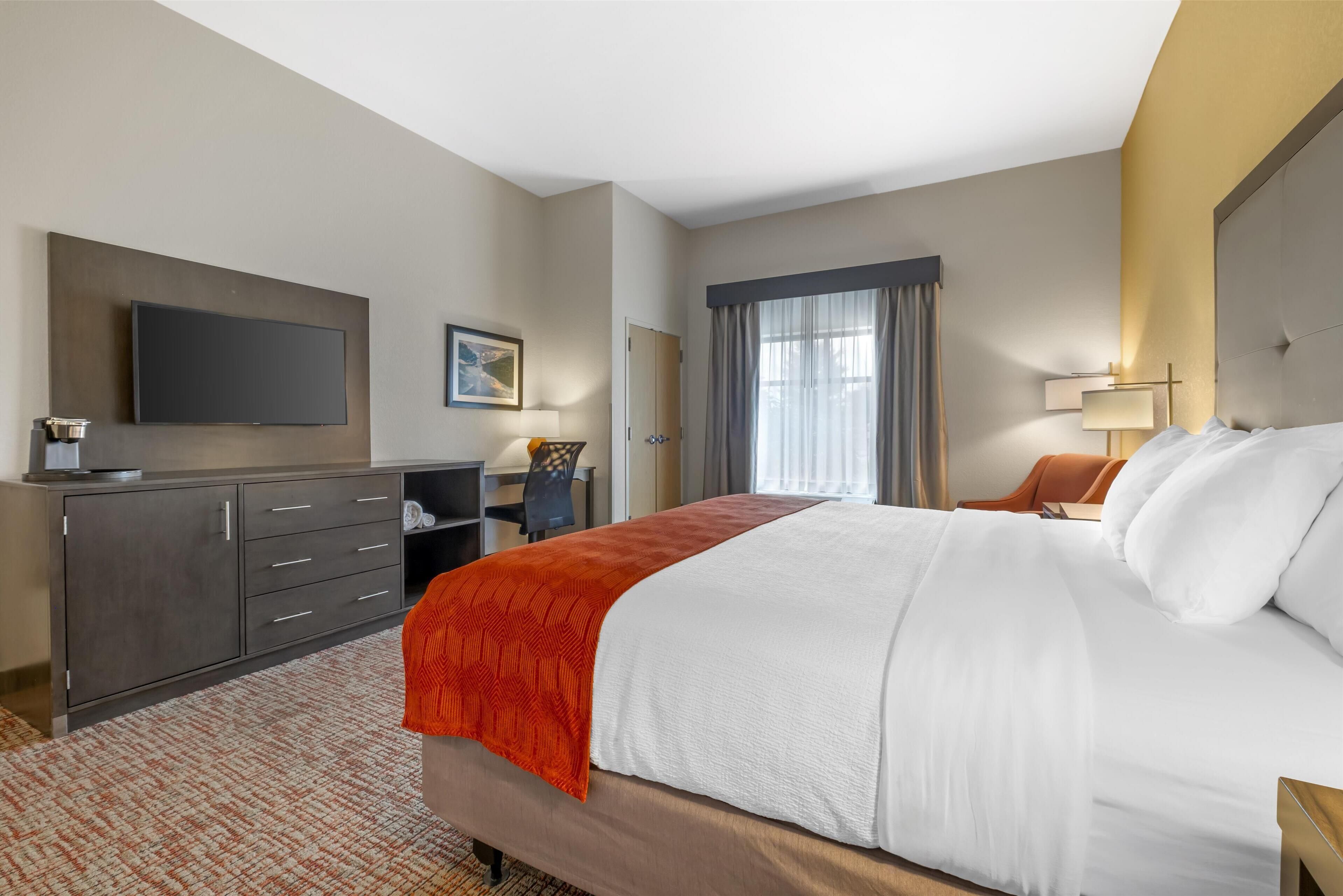 A hotel room with a large bed, a flat-screen TV mounted on the wall, a desk with a chair, a window with curtains, and a bedside table with a lamp.