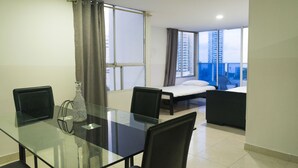 Hotelkamer met een glazen eettafel, twee zwarte stoelen, een bed met een wit matras en een groot raam met gordijnen.