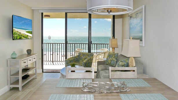 A living room with a view of the ocean, a dining table, and a TV on the wall. - Ideally Situated Beachfront Condo With Pool and Views (Marco Island)