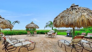 A patio with lounge chairs, a table, and thatched umbrellas. - Tradewinds 1003 (Marco Island)