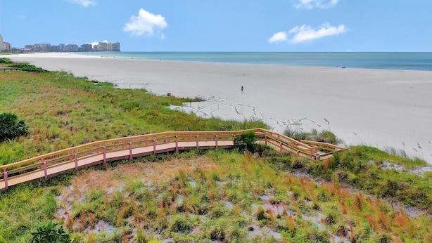 A beach with a wooden boardwalk leading to the shore, a sandy beach, and a distant city skyline. - Tradewinds 1003 (Marco Island)
