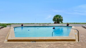 A rectangular swimming pool with a tiled deck, surrounded by palm trees and a view of the beach. - Tradewinds 1003 (Marco Island)
