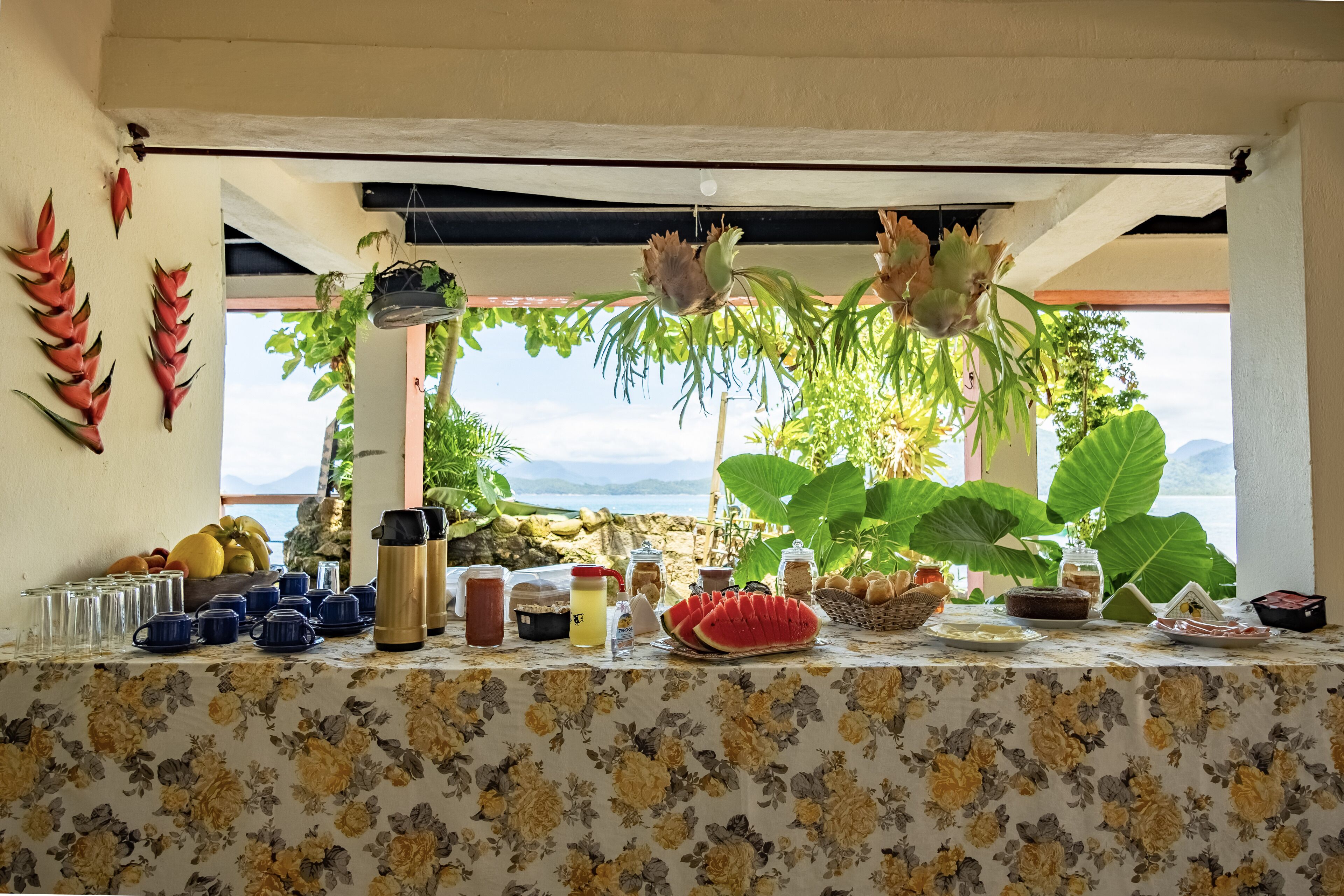 Una mesa con diversos alimentos y bebidas, rodeada de plantas tropicales y una vista panorámica.