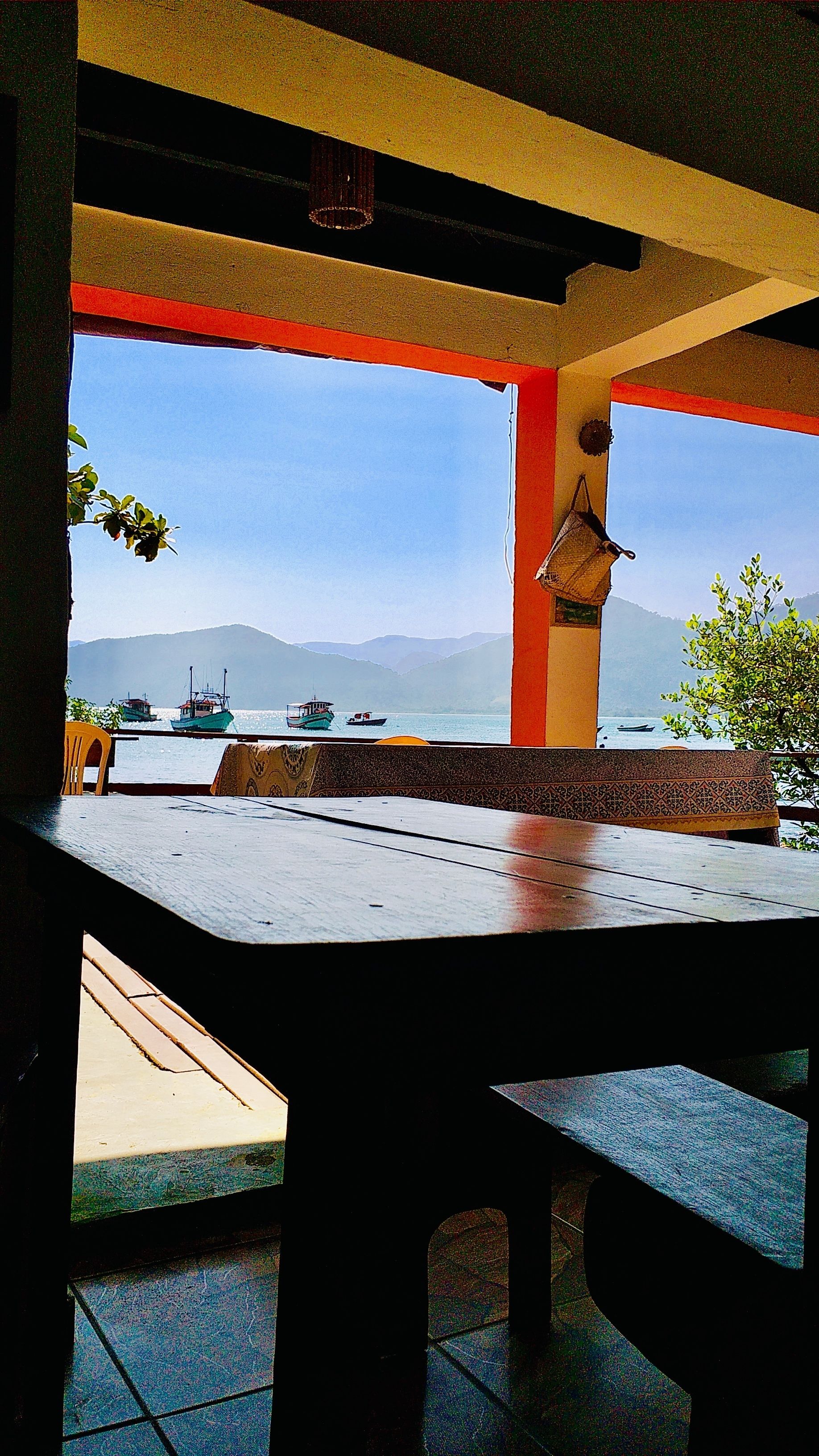 Une vue depuis un espace extérieur abrité, avec une table et des chaises, donnant sur une étendue d’eau où l’on aperçoit des bateaux et, au loin, des montagnes.