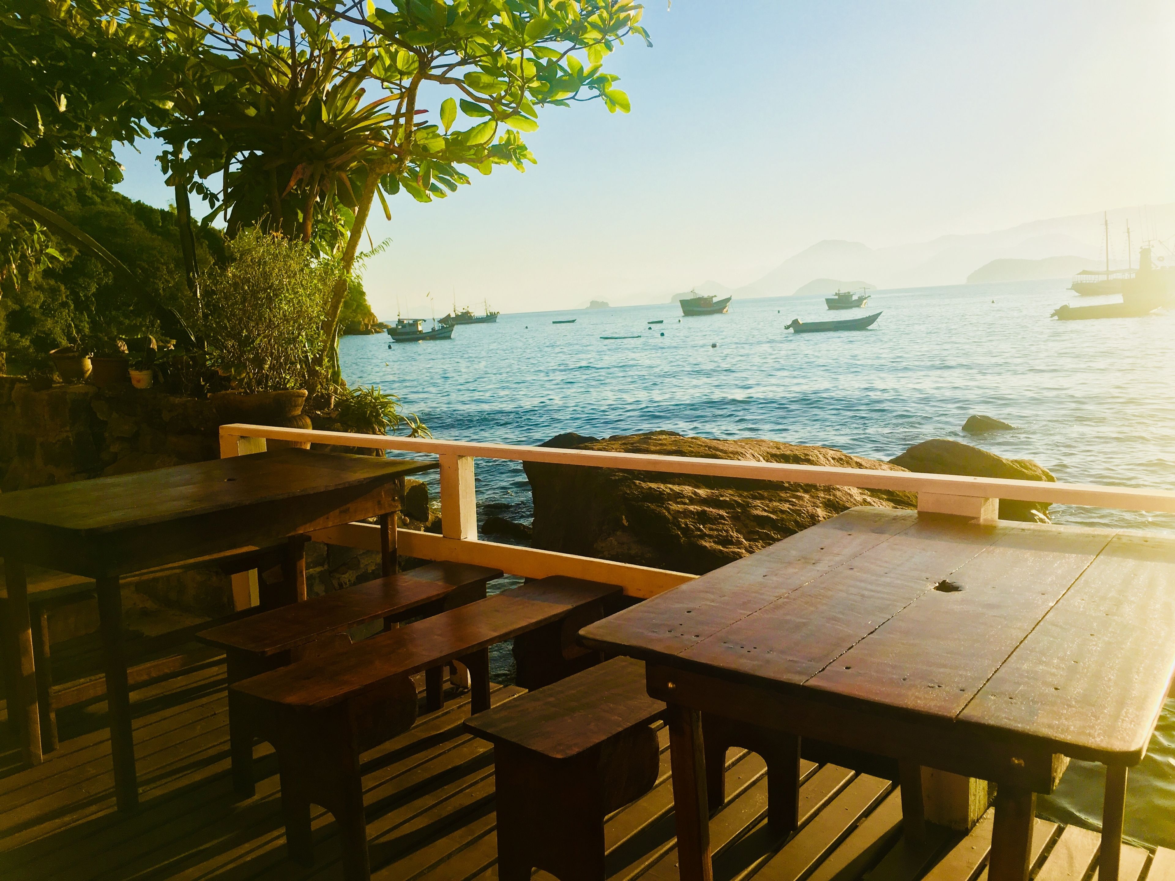 Um deck de madeira com mesas e bancos oferece vista para um mar calmo com barcos.