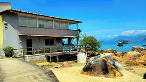 Un edificio costero con balcón que da al mar, muelle de madera y terreno rocoso.