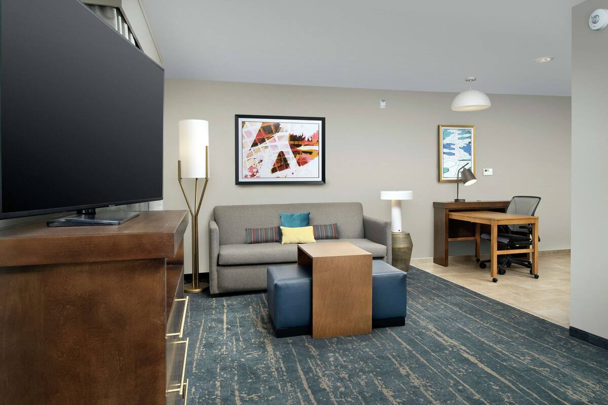 Ett modernt hotellrum med en platt-TV, en soffa, ett soffbord, ett skrivbord med en stol och konstverk på väggarna.