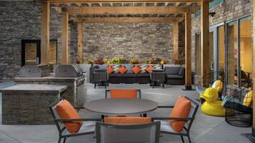 An outdoor patio with stone walls, a wooden pergola, a barbecue area, and outdoor seating with orange cushions.