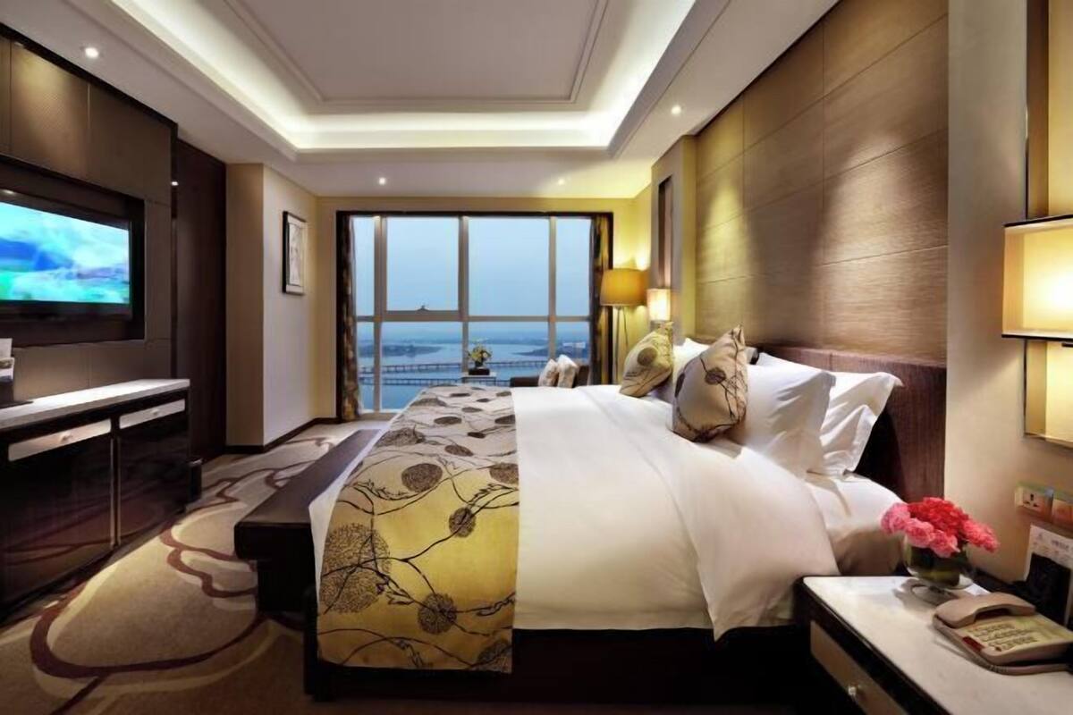 Quarto de hotel com uma cama grande, uma escrivaninha com televisão, vista da cidade e mesa de cabeceira com abajur.