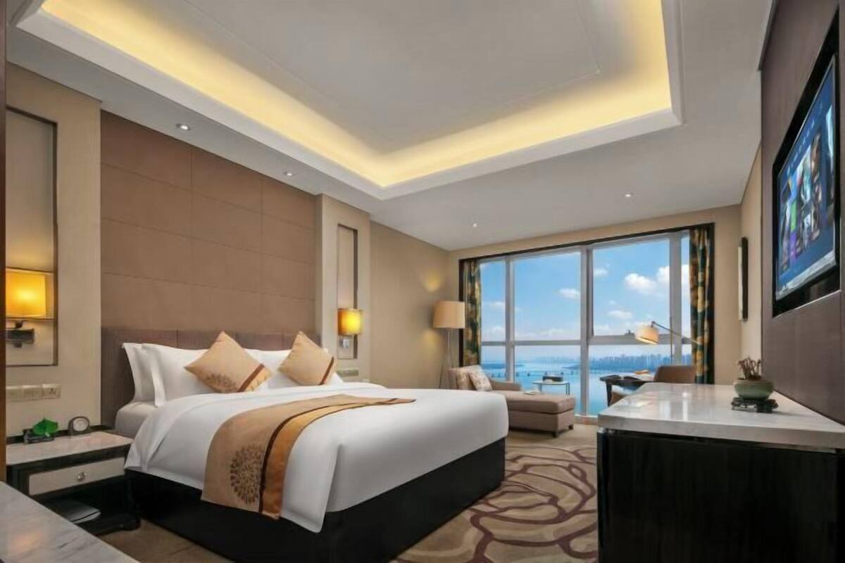 Quarto de hotel com uma cama grande, uma mesa, uma cadeira, vista para o mar e uma janela ampla.