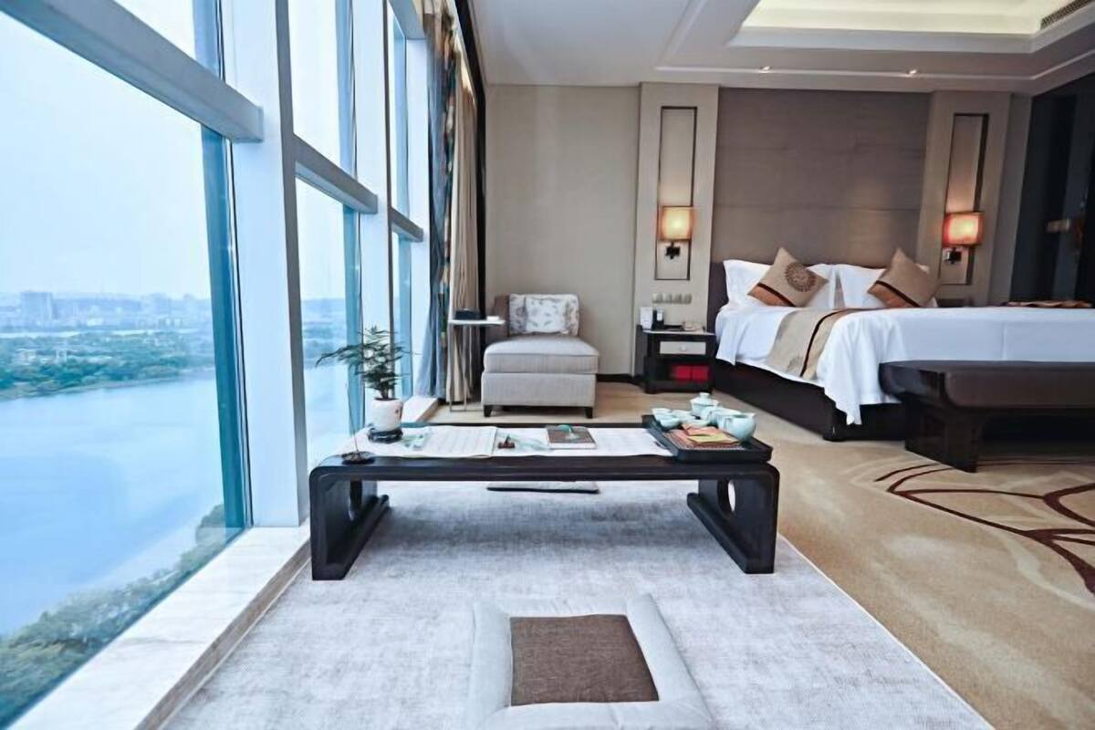 Quarto de hotel com uma janela grande, uma cama, uma mesa de cabeceira e um criado-mudo.