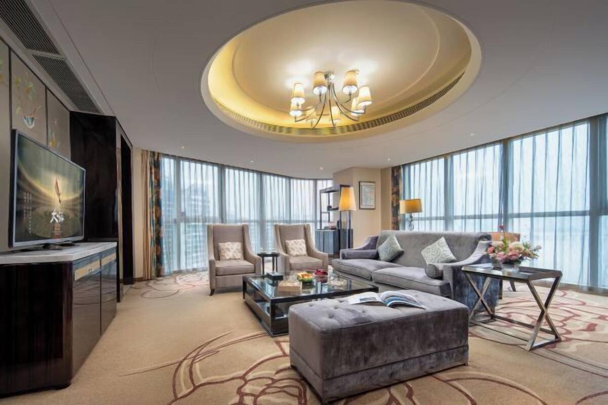 Quarto de hotel moderno com uma televisão grande, assentos confortáveis, uma luminária de teto redonda e janelas amplas.