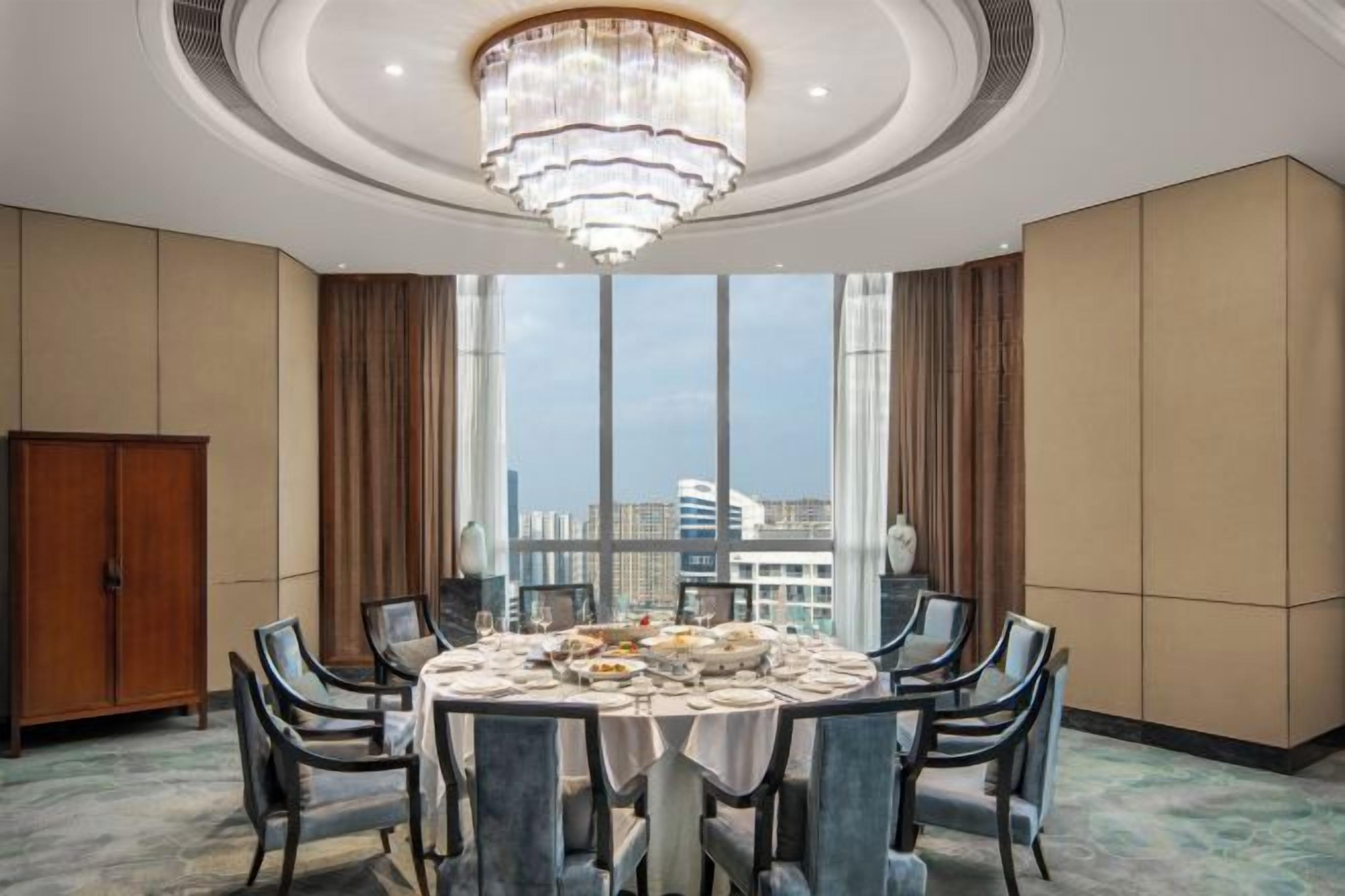 A hotel dining room with a round table, chairs, a chandelier, and large windows offering a city view.