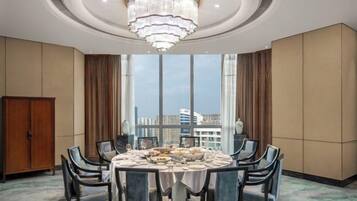 A hotel dining room with a round table, chairs, a chandelier, and large windows offering a city view.