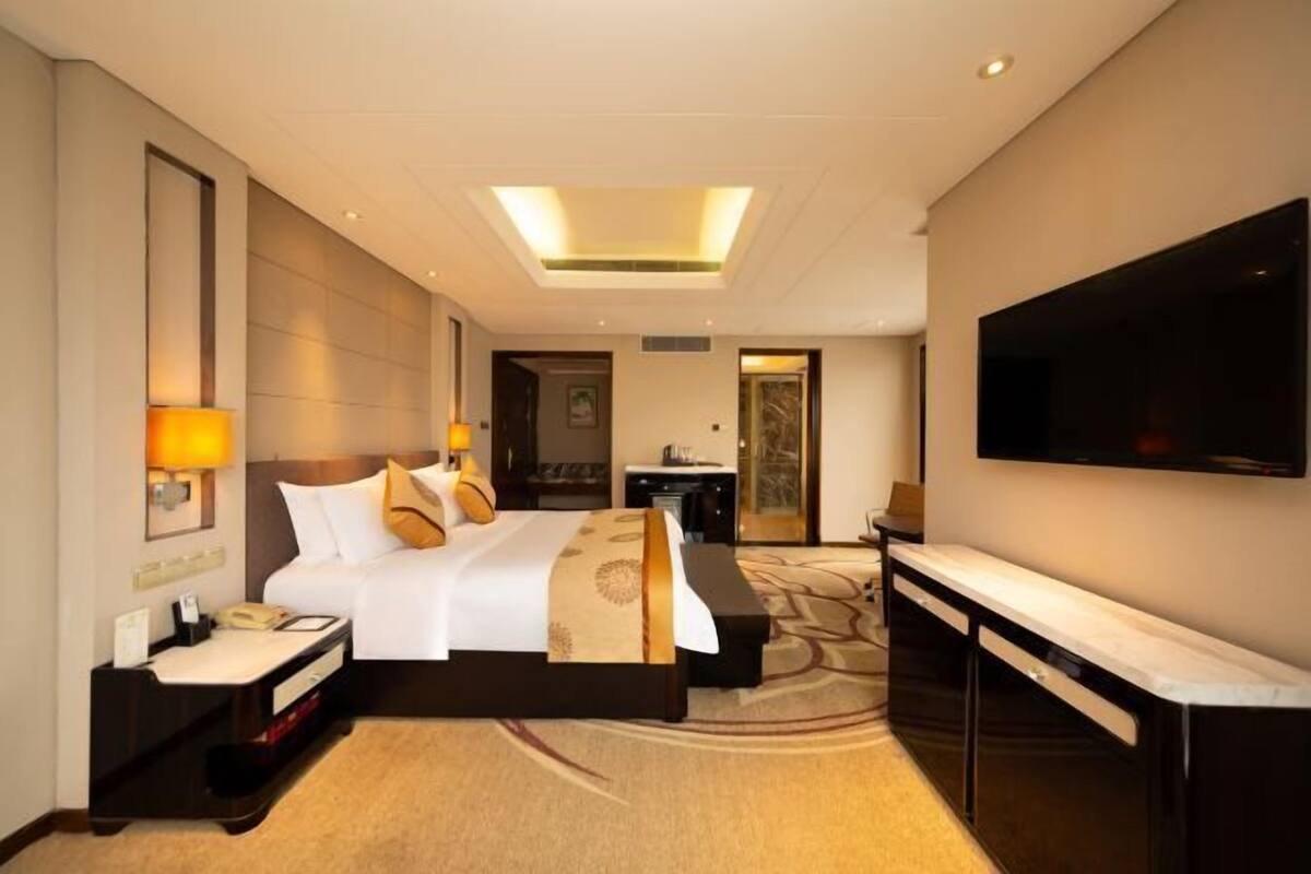 Quarto de hotel com uma cama grande, uma escrivaninha com telefone, uma TV de tela plana na parede e piso coberto de carpete.