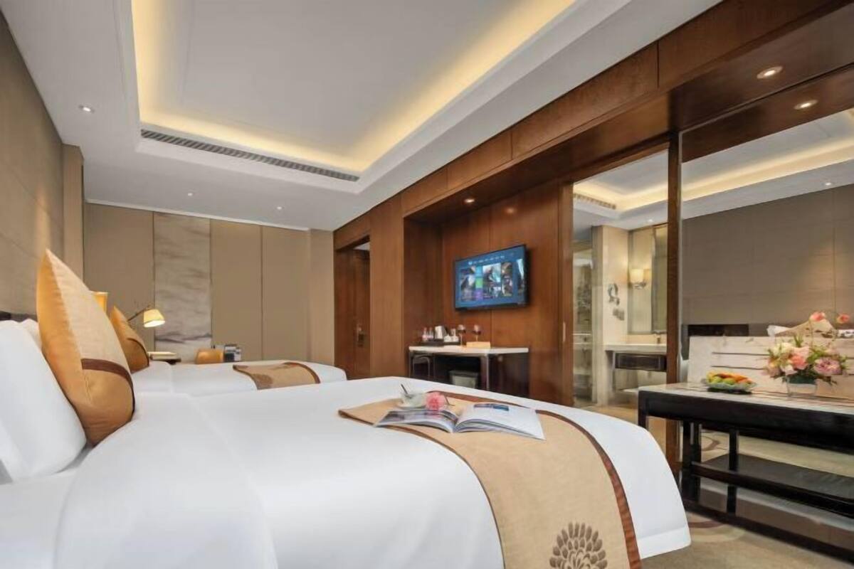 Quarto de hotel com uma cama grande, uma escrivaninha com computador, televisão e um banheiro visível através de um painel de vidro.