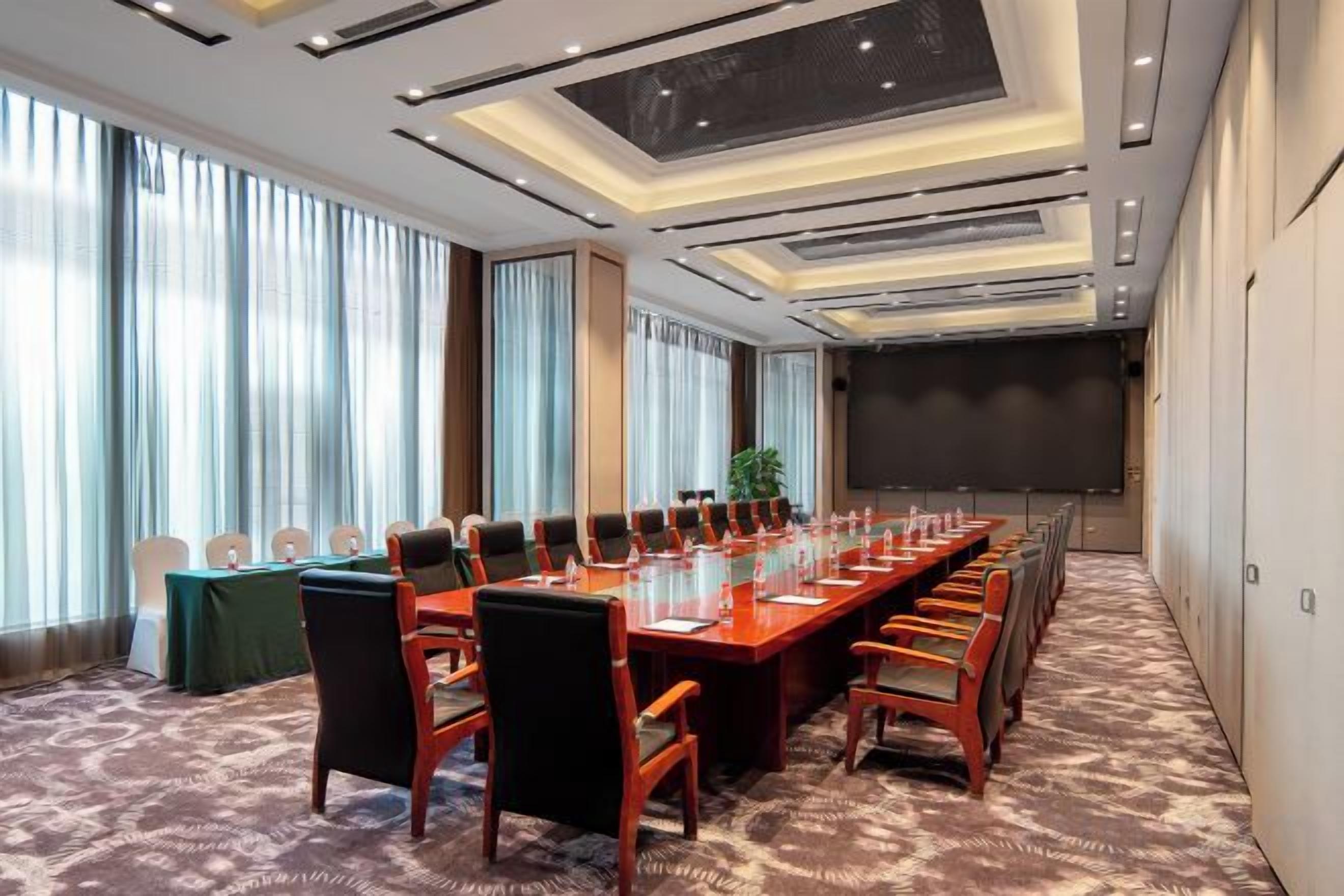 Un salón de conferencias con una larga mesa, sillas, ventanas amplias y una pantalla de proyector.
