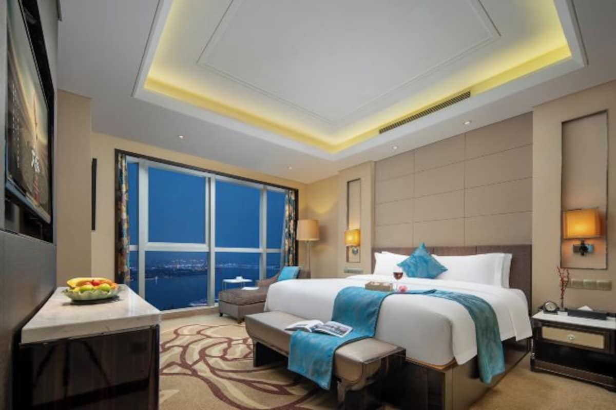 A hotel room with a large bed, a desk with a fruit bowl, a view of the city, and a modern ceiling design.