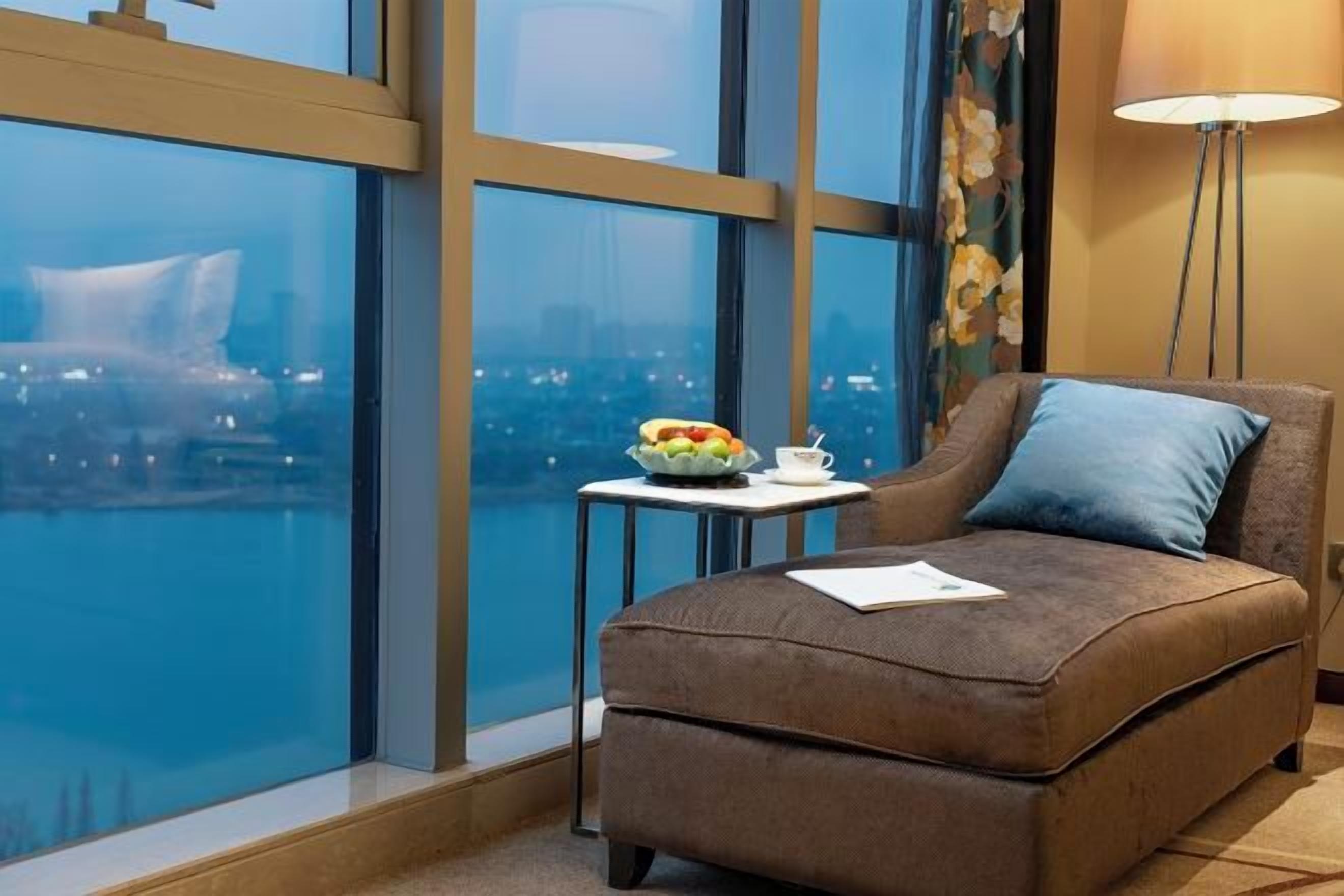 A cozy room with a large window, a small table with a bowl of fruit, and a cushioned seating area.