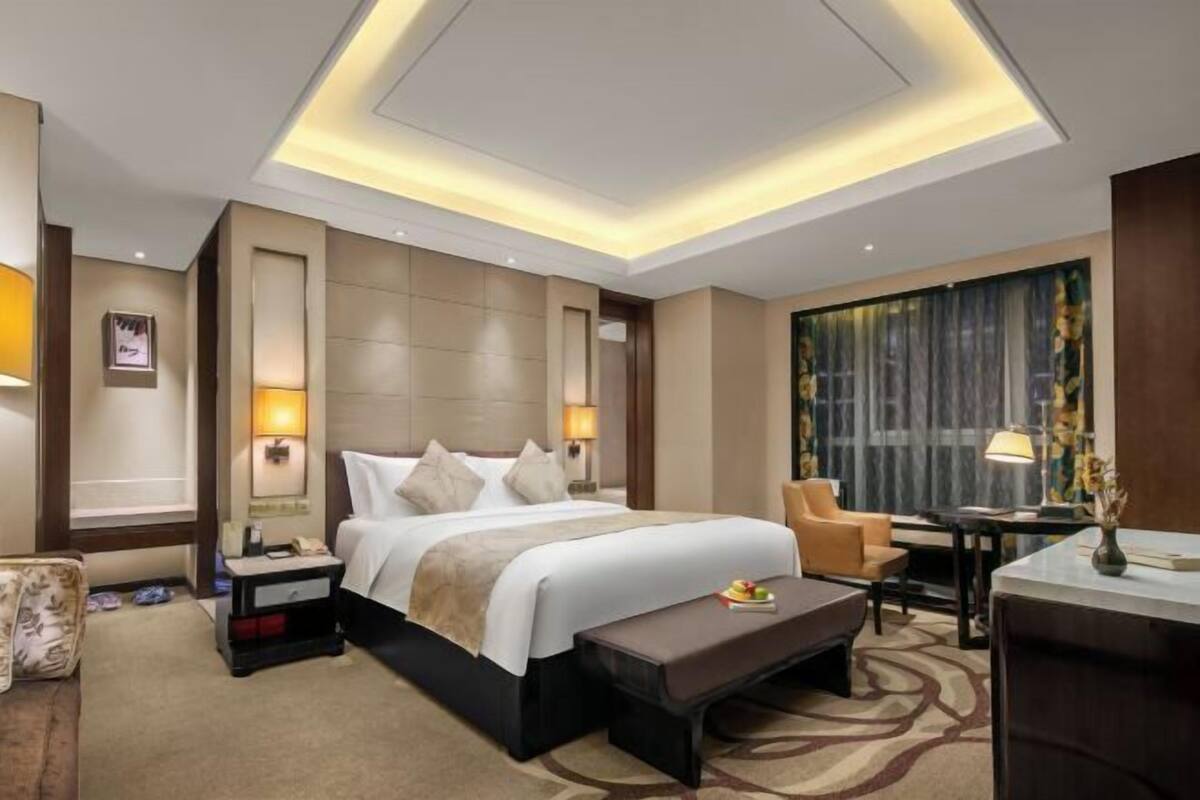 Quarto de hotel com cama, mesas de cabeceira, escrivaninha, uma cadeira e uma janela grande com cortinas.