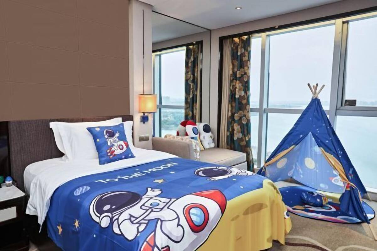 Quarto de hotel com uma cama e um edredom com tema espacial, uma janela com vista e um tipi azul.