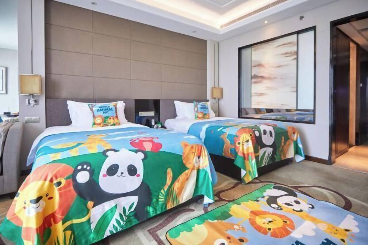 Quarto de hotel com duas camas, cada uma com uma colcha colorida com tema de animais, uma janela grande com vista e uma cabeceira moderna.