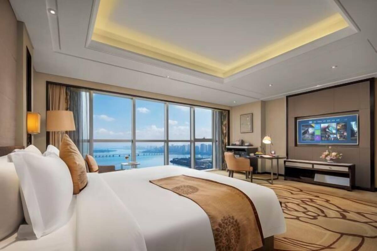 Quarto de hotel com uma cama grande, uma escrivaninha, uma televisão e vista para a paisagem urbana.