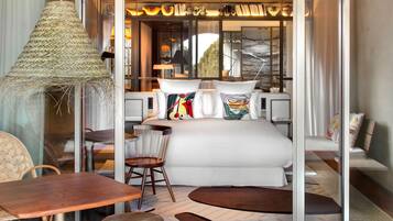 Una habitación de hotel moderna con una cama grande, una zona de comedor con mesa y sillas, una lámpara de fibras naturales y vistas al exterior a través de amplios ventanales.