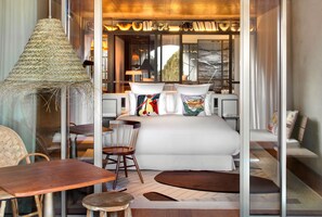 Una habitación de hotel moderna con una cama grande, una zona de comedor con mesa y sillas, una lámpara de fibras naturales y vistas al exterior a través de amplios ventanales.