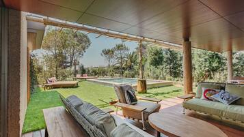 An outdoor patio with a pool, wooden furniture, and a view of the surrounding greenery.