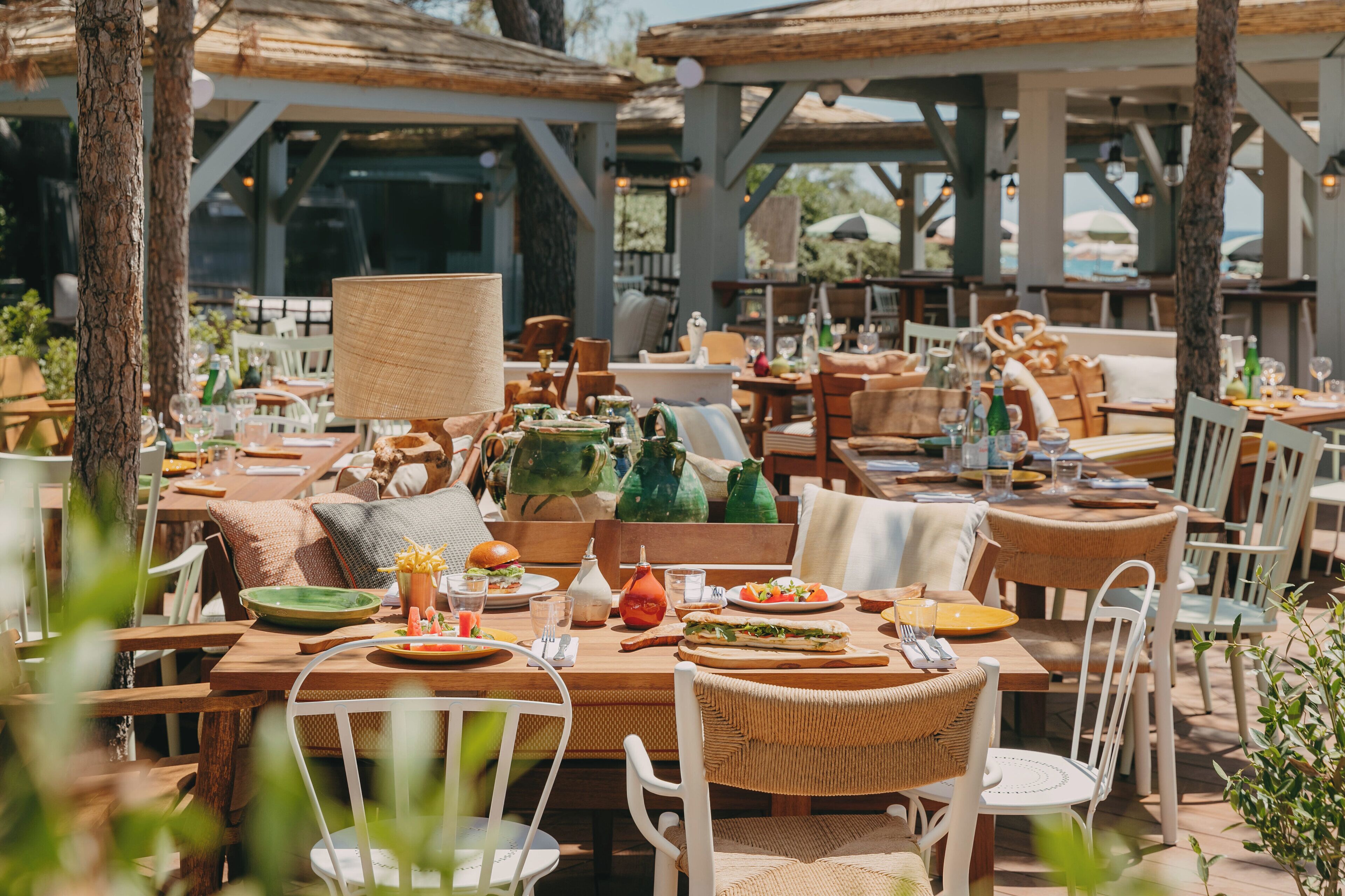 Un área de comedor al aire libre con mesas y sillas de madera, rodeada de árboles y cubierta por pérgolas de madera.