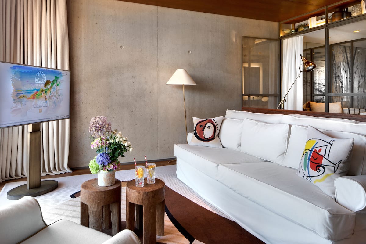 Un salon moderne avec un canapé blanc, une table basse en bois ornée d’un vase de fleurs et un téléviseur à écran plat placé sur le côté gauche.