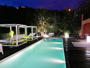 Une piscine avec des chaises longues et un espace de repos couvert, entourée d’arbres et avec un bâtiment en arrière-plan.
