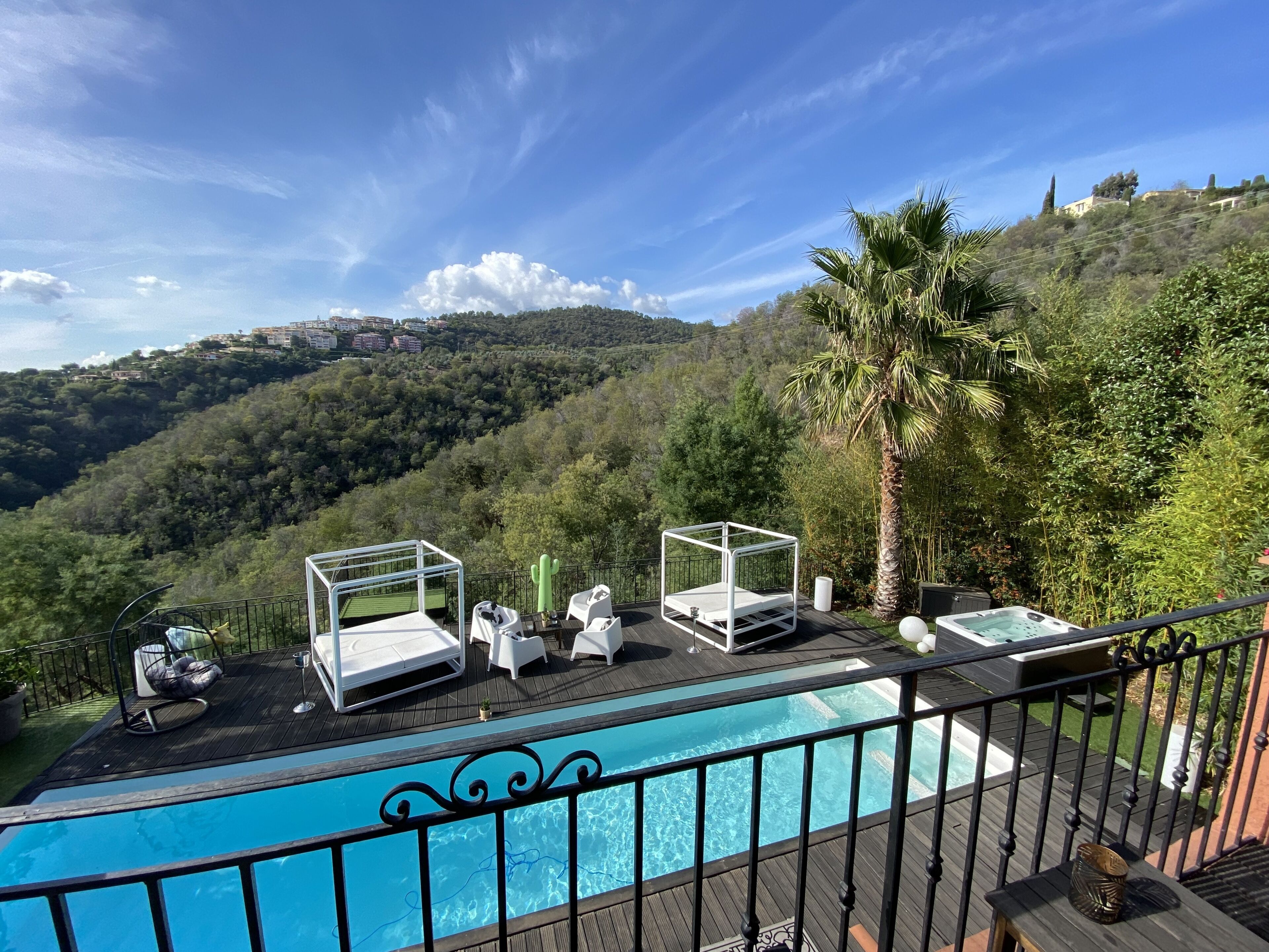 A poolside area with two white sunbeds and a hot tub, surrounded by lush greenery and a view of a hillside.
