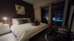 A hotel room with a bed, a nightstand, a chair, a window with a view of the city at night, and a painting on the wall. - Villa Colina (Mandelieu-La-Napoule)