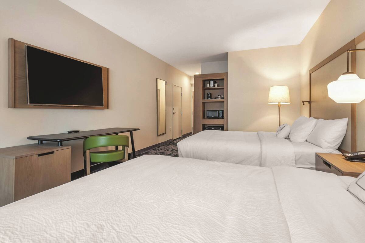 A hotel room with two beds, a desk, a chair, a TV, a lamp, and a shelf with toiletries.