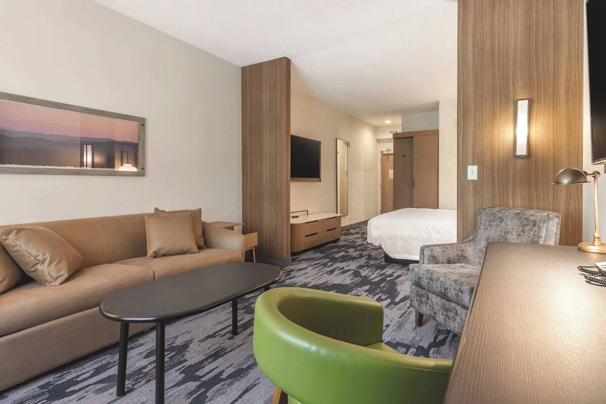 A hotel room with a sofa, a green armchair, a bed, a desk, and a television.