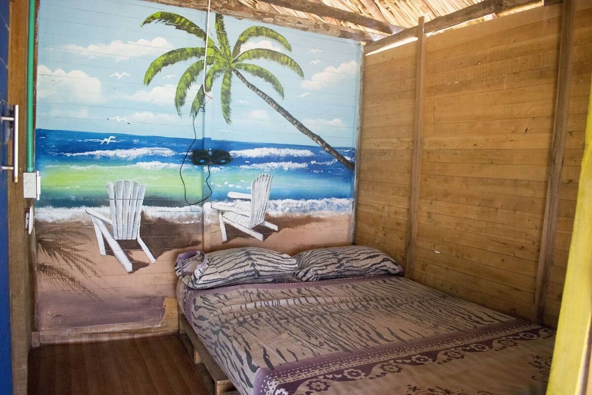 A room with a mural of a beach scene, a bed with patterned bedding, and two white chairs.
